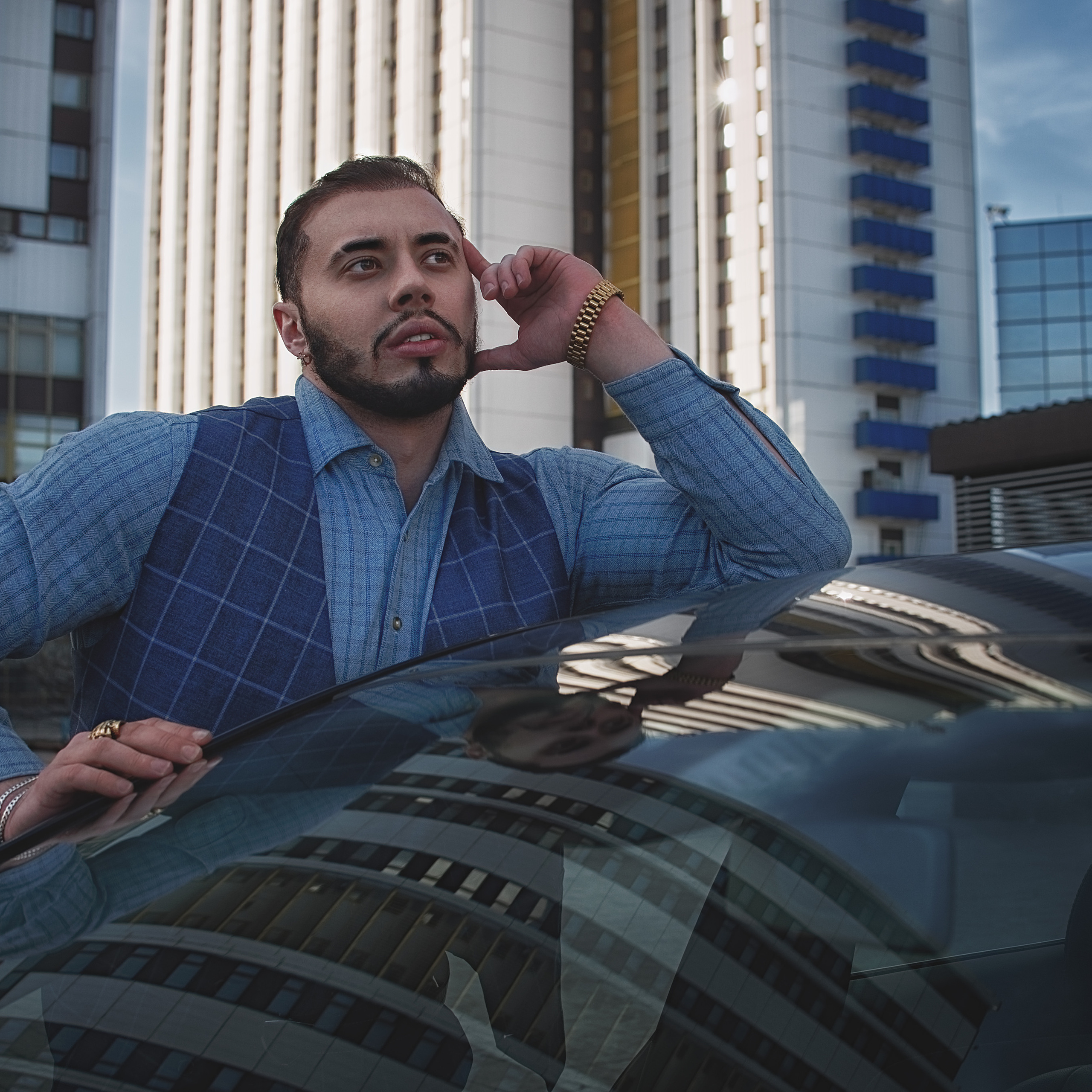 Задумчивый мужчина стоит возле машины, на заднем фоне здания