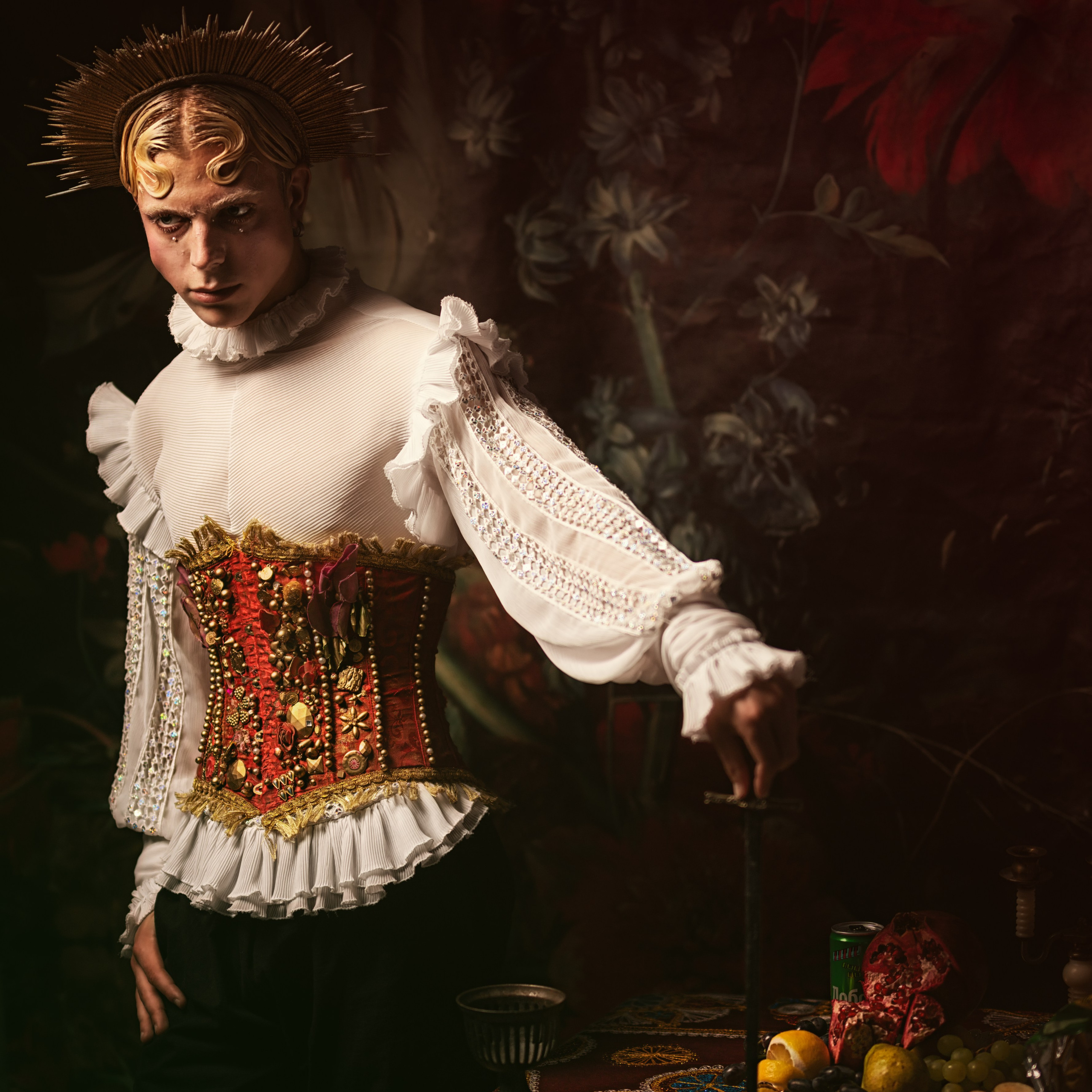 человек в костюме эпохи Возрождения. На нём белый корсет с пышными рукавами, украшенный золотыми и красными элементами, а также изысканный головной убор, напоминающий корону. Фон выполнен в тёмных тонах с цветочными мотивами, что подчёркивает драматичность образа. В руках человек держит длинный тонкий предмет, а на столе перед ним расположены фрукты и другие декоративные элементы.Заголовок: «Королевский стиль эпохи Возрождения»Ключевые слова: исторические костюмы, эпоха Возрождения, корсеты, викторианский стиль, портретный фотограф, Иван Сафронов.
