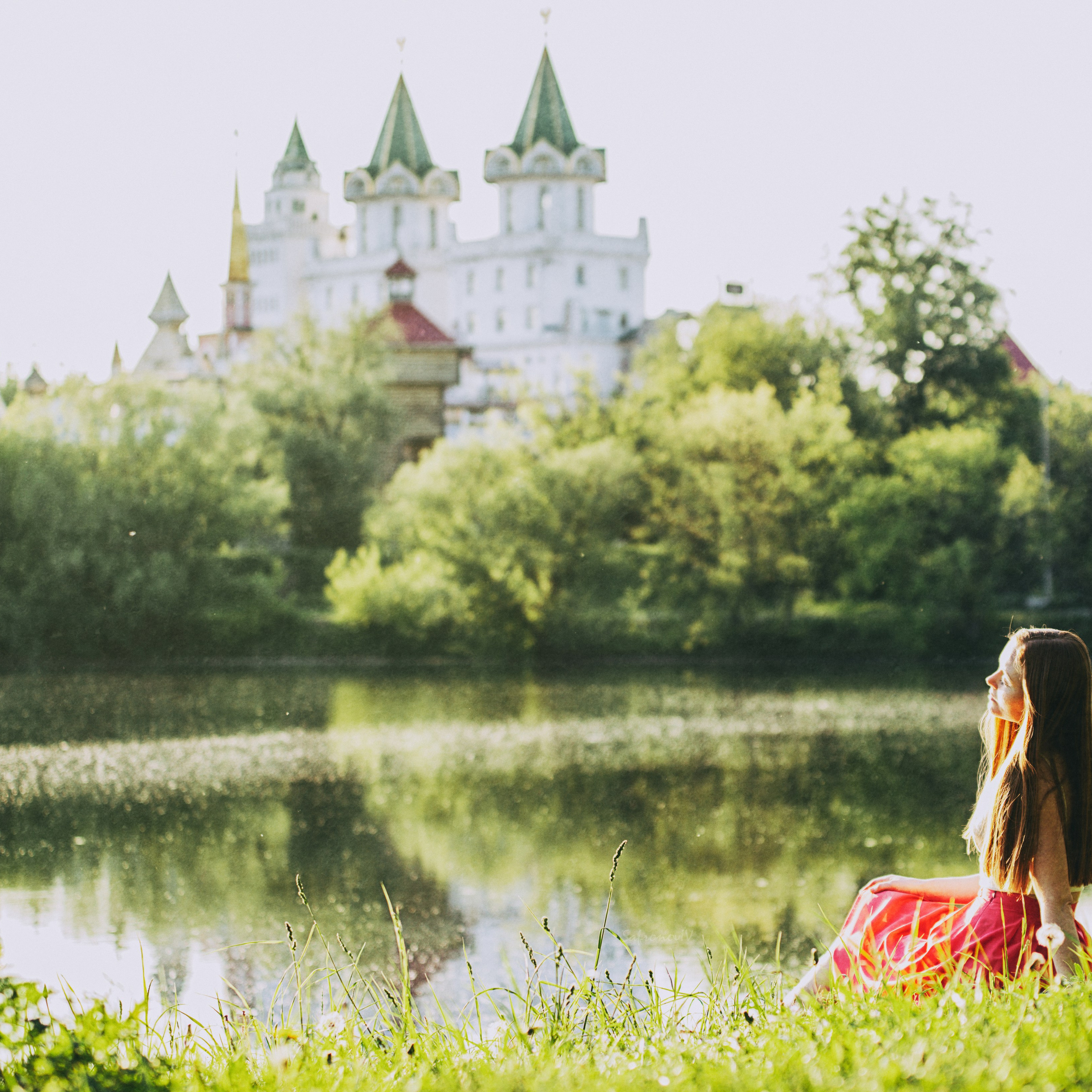 девушка в ярком красном платье, сидящая на траве у воды. На заднем плане виден Измайловский кремль, окружённый зелёными деревьями. Атмосфера снимка спокойная и романтичная, подчёркивающая гармонию природы и архитектуры.Заголовок: «Мечтательная красота у воды»Ключевые слова: фотосессия, природа, вода, кремль, девушка, красное платье, лето, Москва, фотограф Сафронов Иван.