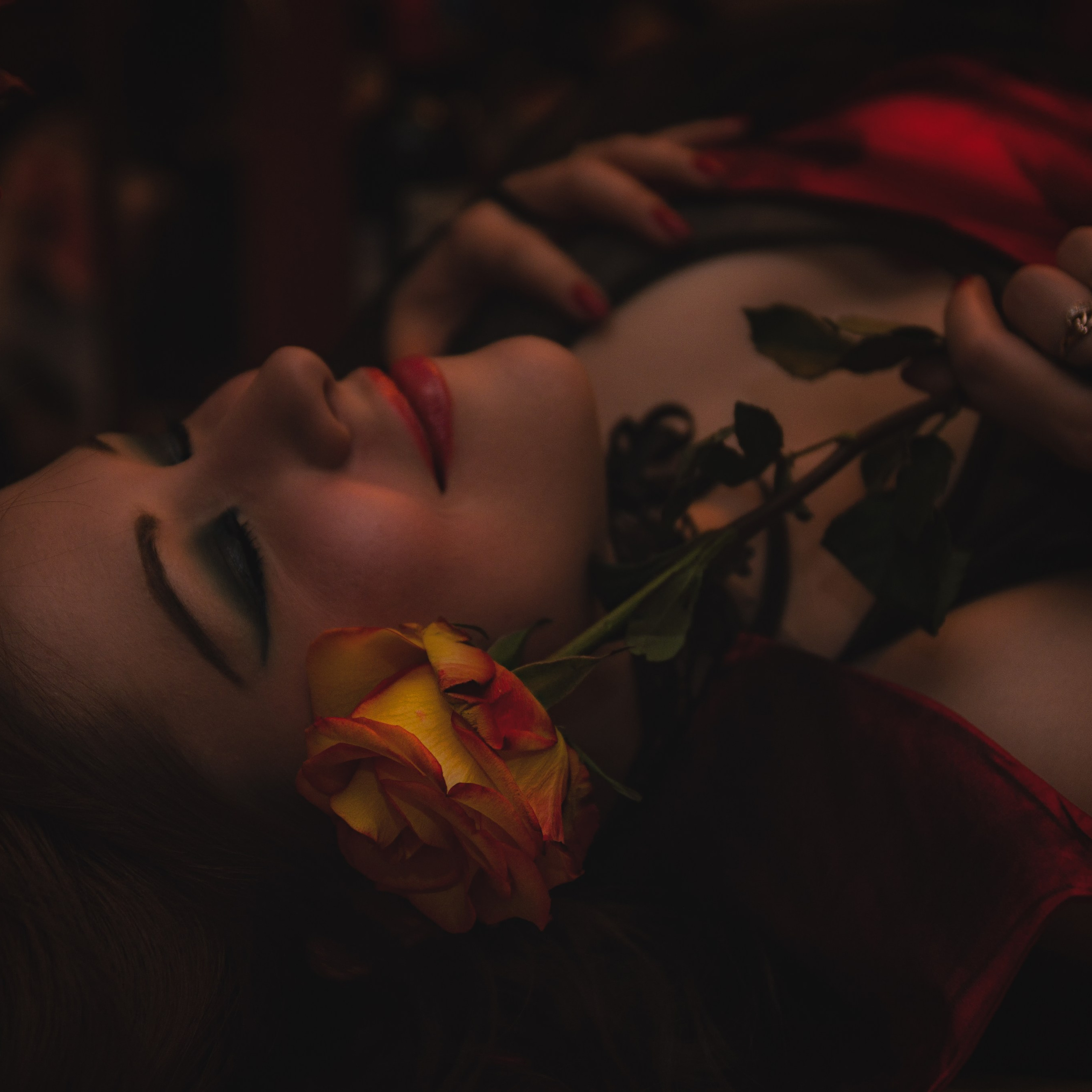 женщина с закрытыми глазами, лежащая на боку. Её лицо обрамлено красно-жёлтой розой, которая добавляет образу романтики и загадочности. Женщина одета в красное платье, её макияж подчёркивает выразительность глаз и губ. Атмосфера снимка наполнена теплом и интимностью, а мягкий свет акцентирует внимание на деталях.Заголовок: «Тайна розы»Ключевые слова: фотосессия с розами, женщина с розами, романтическая фотосессия, портретный фотограф, Иван Сафронов.