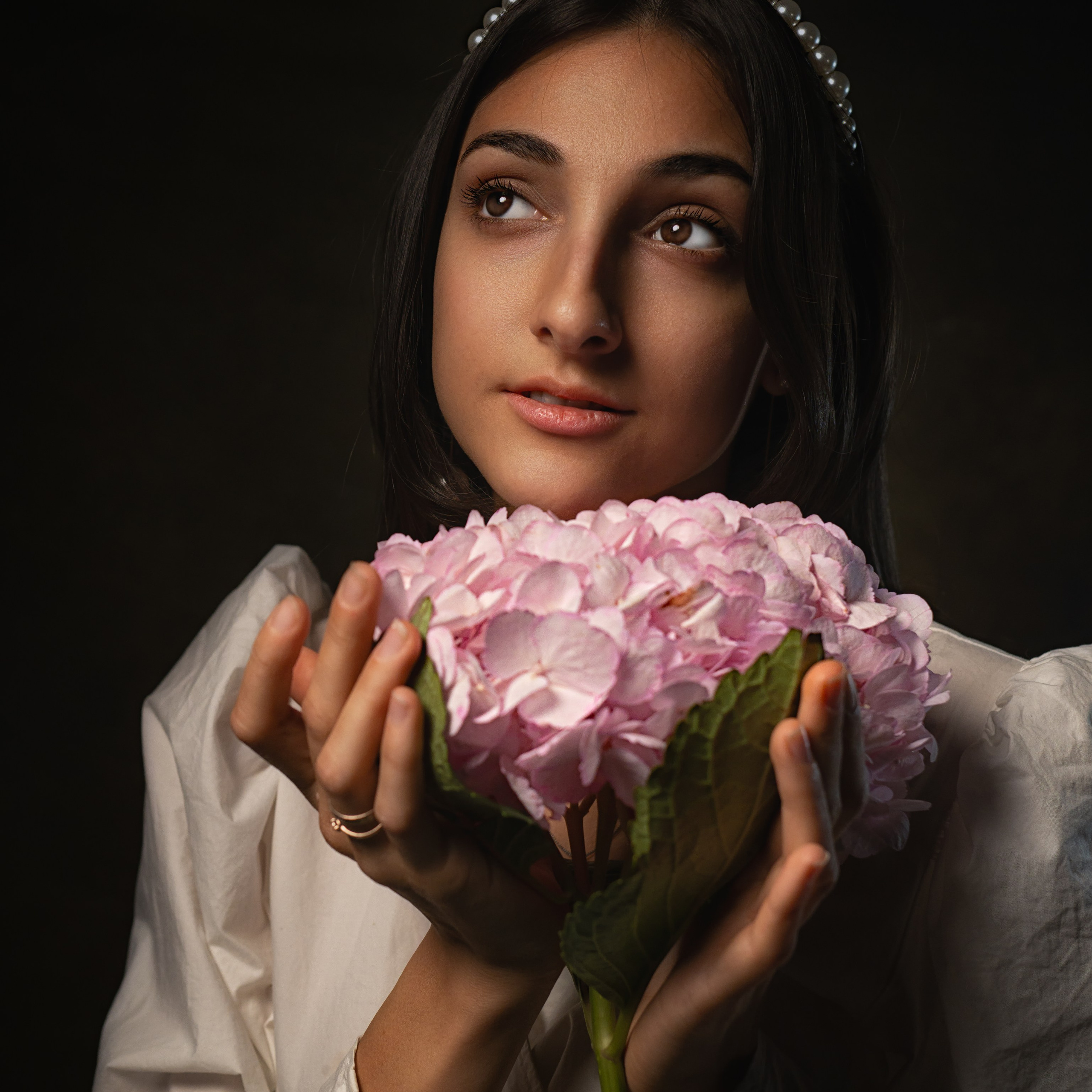 молодая женщина с темными волосами, украшенными жемчужным ободком. Она держит в руках большой розовый цветок гортензии, который занимает центральное место в композиции. Фон темный, что подчеркивает мягкость света, падающего на лицо и руки модели. Атмосфера снимка романтичная и нежная, с акцентом на естественную красоту и гармонию.Заголовок: «Гармония природы и красоты»Ключевые слова: портрет, женщина, гортензия, жемчужный ободок, романтический стиль, портретный фотограф, Москва, Сафронов Иван.