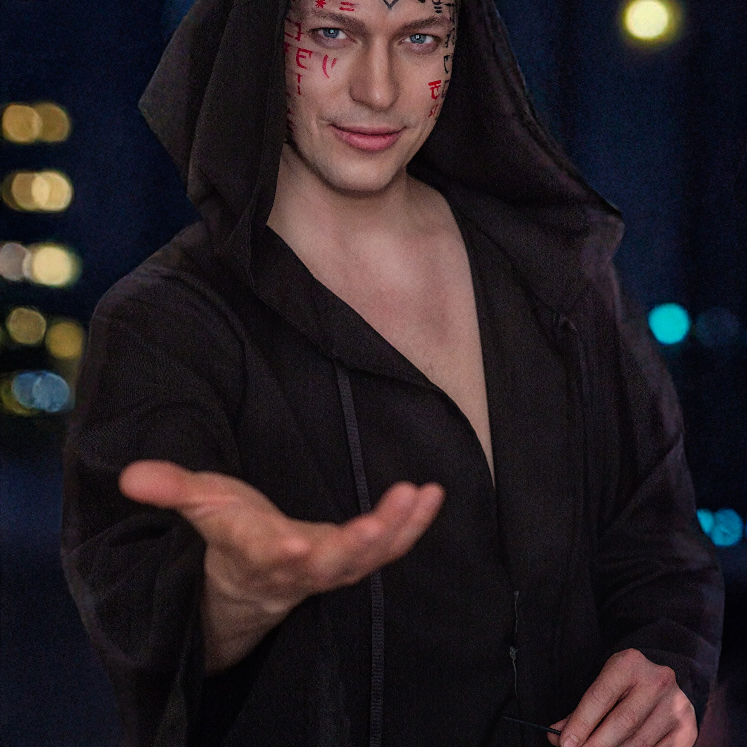 На фотографии изображён мужчина в чёрной одежде с капюшоном, который слегка откинут назад. Его лицо украшает яркий макияж с красными и чёрными элементами, создающими загадочный и мистический образ. В руках он держит солнцезащитные очки, а его взгляд направлен прямо в камеру, создавая ощущение интриги. Фон размытый, с яркими огнями, что добавляет глубину и атмосферу снимку.Заголовок: «Мистический маг в городе»Ключевые слова: экстрасенс, магия, мистика, макияж, очки, портрет, фотограф, Сафронов Иван, Москва.