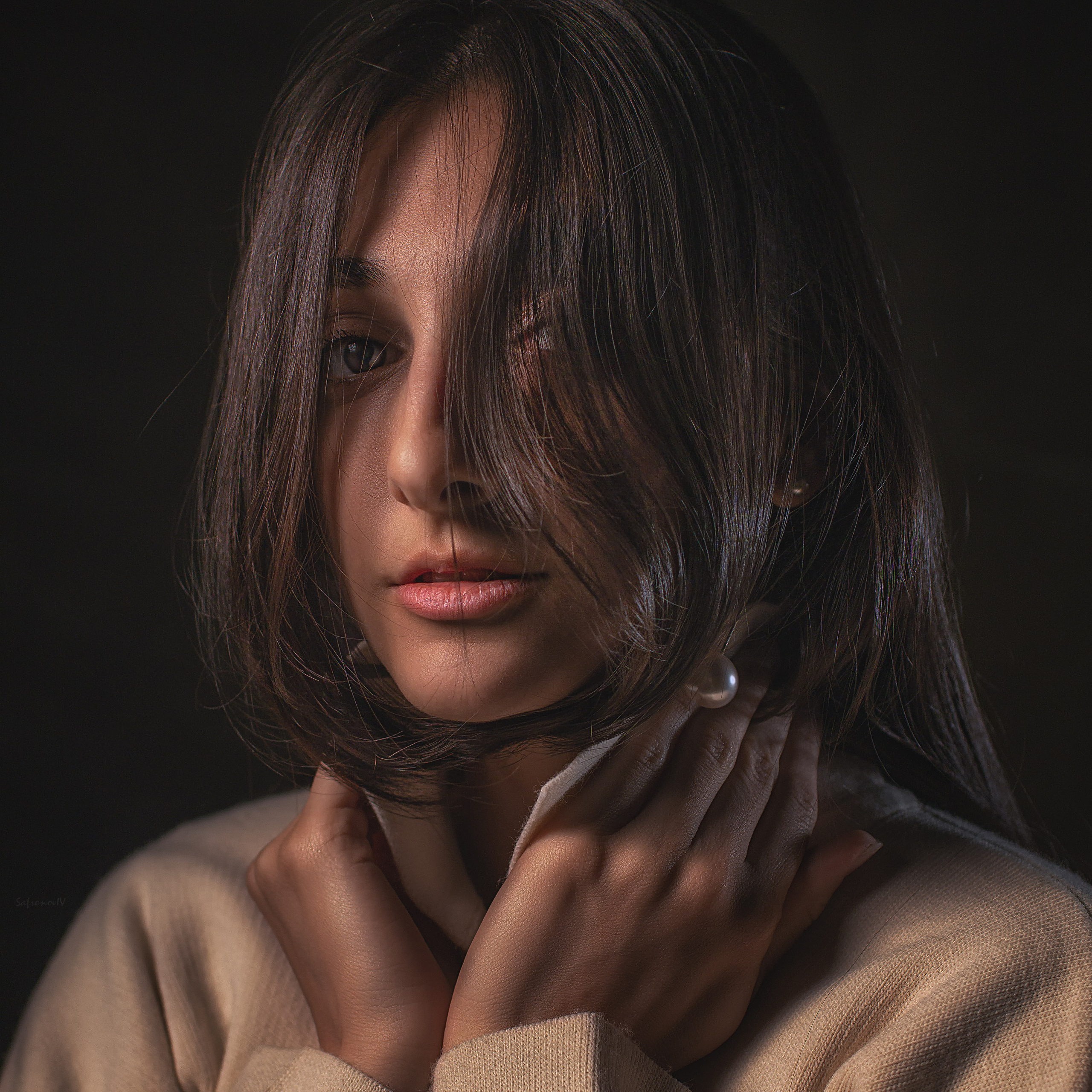 молодая женщина с длинными темными волосами, которые частично закрывают лицо. Она одета в светлый свитер, а в ухе видна жемчужная серьга. Взгляд направлен в сторону, создавая ощущение задумчивости и легкой меланхолии. Освещение мягкое, с акцентом на лице, что подчеркивает естественную красоту модели. Фон темный, что добавляет глубину и драматизм снимку.Заголовок: «Загадочный взгляд: портрет в стиле минимализма»Ключевые слова: портрет, девушка, длинные волосы, жемчужная серьга, минимализм, мягкое освещение, задумчивость, драматический фон, фотограф Сафронов Иван, портретная съемка