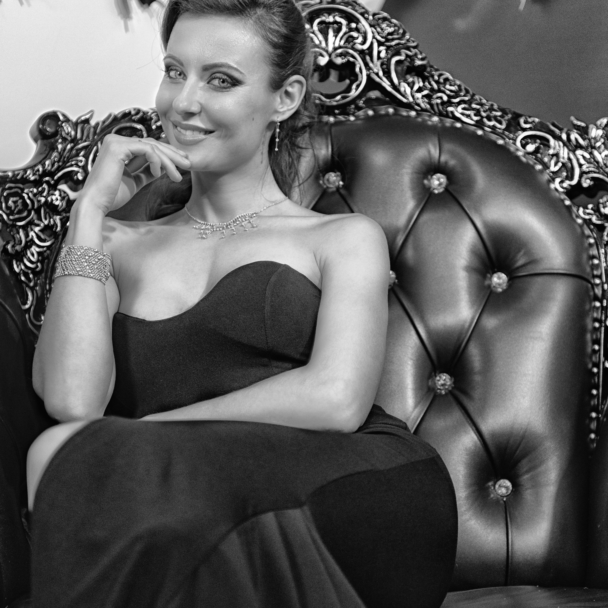 элегантная женщина в черном платье, сидящая на роскошном кожаном кресле с резной спинкой. Ее образ дополняют ювелирные украшения, включая браслет и ожерелье. Фон украшен декоративными элементами, создающими атмосферу утонченности и стиля. Черно-белая гамма подчеркивает изысканность и драматизм снимка.Заголовок: «Элегантность в черно-белом»Ключевые слова: портретный фотограф, Москва, Сафронов Иван, черно-белая фотография, элегантность, стиль, ювелирные украшения, кожаное кресло.