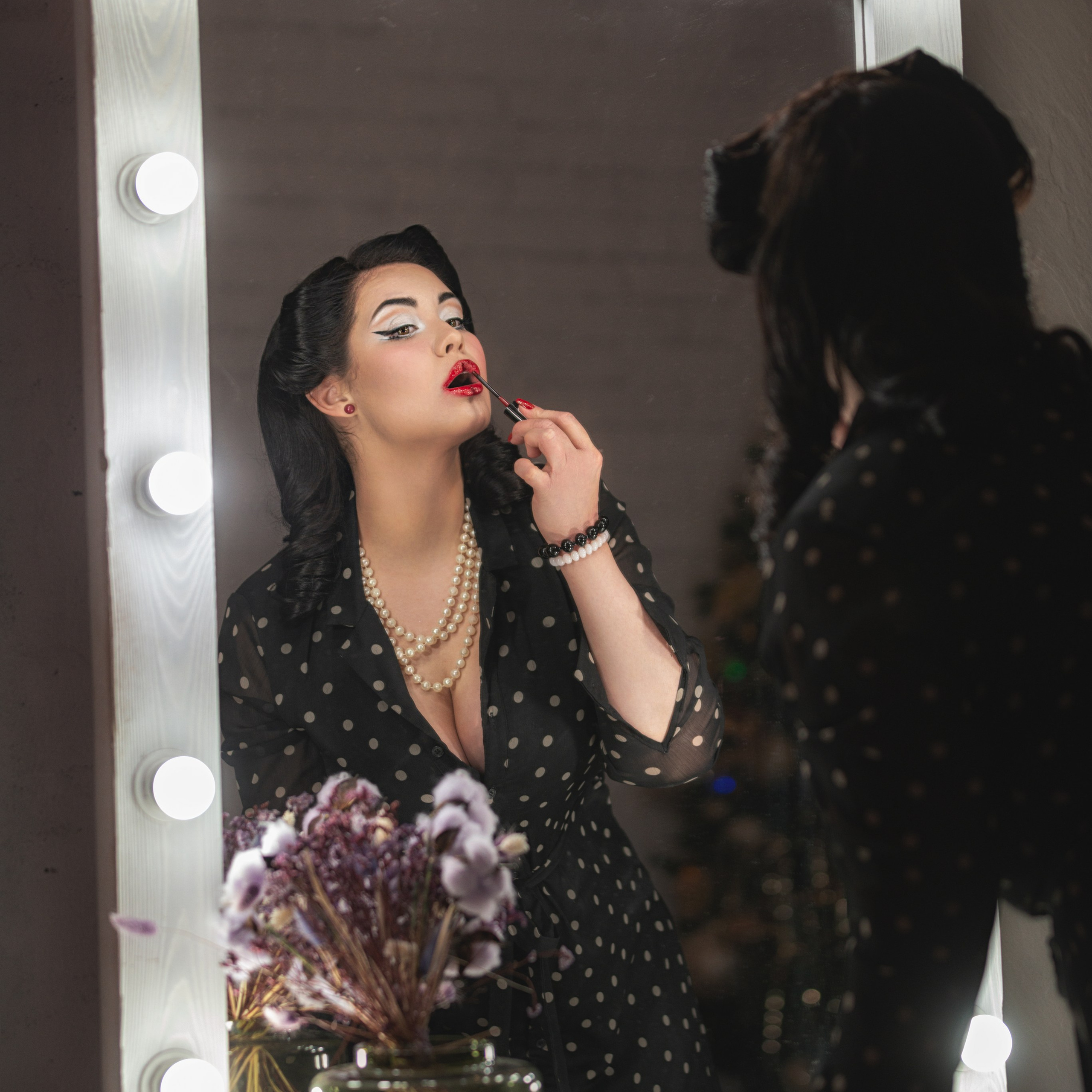 На изображении изображена женщина, готовящаяся к выходу в свет. Она наносит яркую красную помаду перед зеркалом с подсветкой, создавая атмосферу ретро-шика. Её образ дополняют жемчужное ожерелье, элегантное чёрное платье в горошек и изысканная причёска. На столе перед ней стоит стеклянная ваза с сухоцветами, что добавляет уюта и стиля.Заголовок: «Элегантность в деталях: момент преображения»Ключевые слова: портретный фотограф в Москве, фотограф Сафронов Иван, ретро-стиль, макияж, зеркало с подсветкой, жемчужное ожерелье.