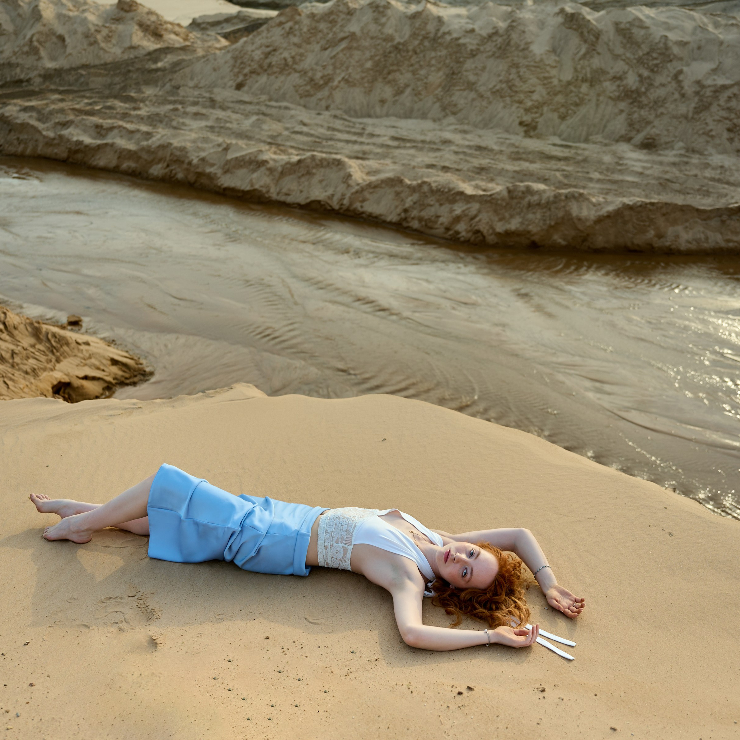 Лежит у кромки воды, ноги согнуты за спиной, тёплый закатный свет