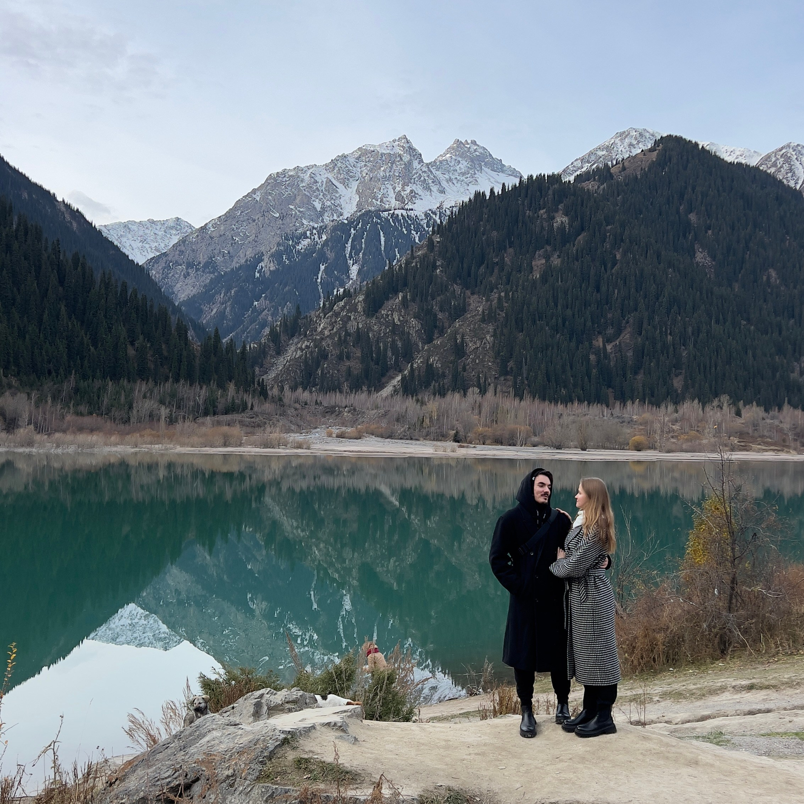 Двое людей стоят на фоне живописного горного озера Иссык, отражающего заснеженные вершины и густые хвойные леса. Озеро спокойное, с ярко-зелёной водой, и создаёт чувство гармонии с природой. Люди на переднем плане выглядят счастливыми, их фигуры контрастируют с величественным пейзажем гор на заднем плане под светло-серым небом.