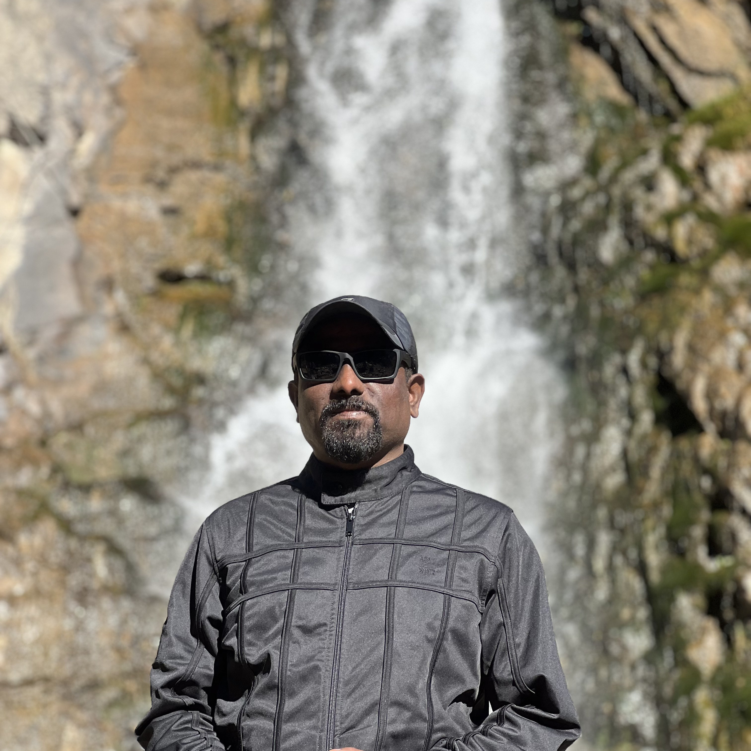 Мужчина в тёмной куртке и солнечных очках стоит на фоне водопада Медвежий в районе Алматы. На нём также надета кепка. В руках он держит телефон красного цвета. За его спиной струи воды спадают с утёса, создавая впечатляющий водный фон.