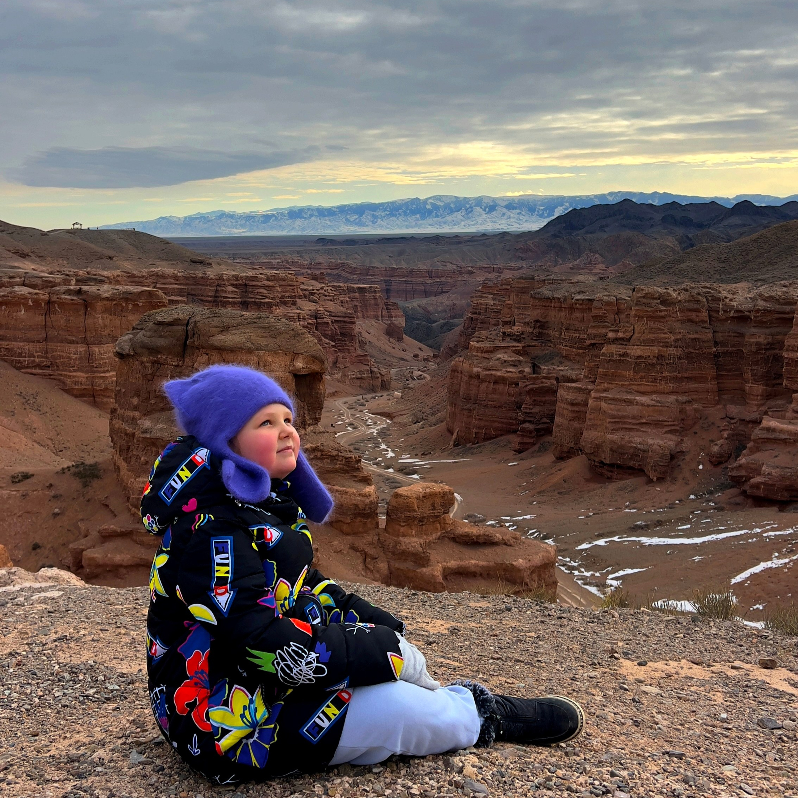 Маленький ребенок в яркой зимней куртке с разноцветными узорами и фиолетовой шапке с ушами сидит на склоне с видом на живописный Чарынский каньон. За ним видны величественные красные скалы, ущелья и долины, частично покрытые снегом. На заднем плане, вдали, можно заметить горный хребет под облачным небом, создающим атмосферу спокойствия и красоты природы.