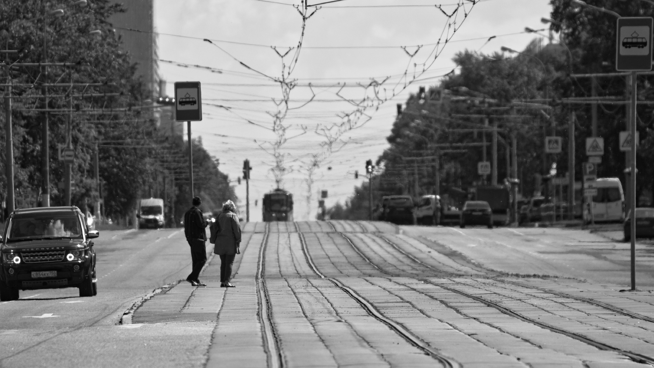 Два человека идут по трамвайным путям под переплетением контактных проводов, трамвай вдали, черно-белое фото городской инфраструктуры