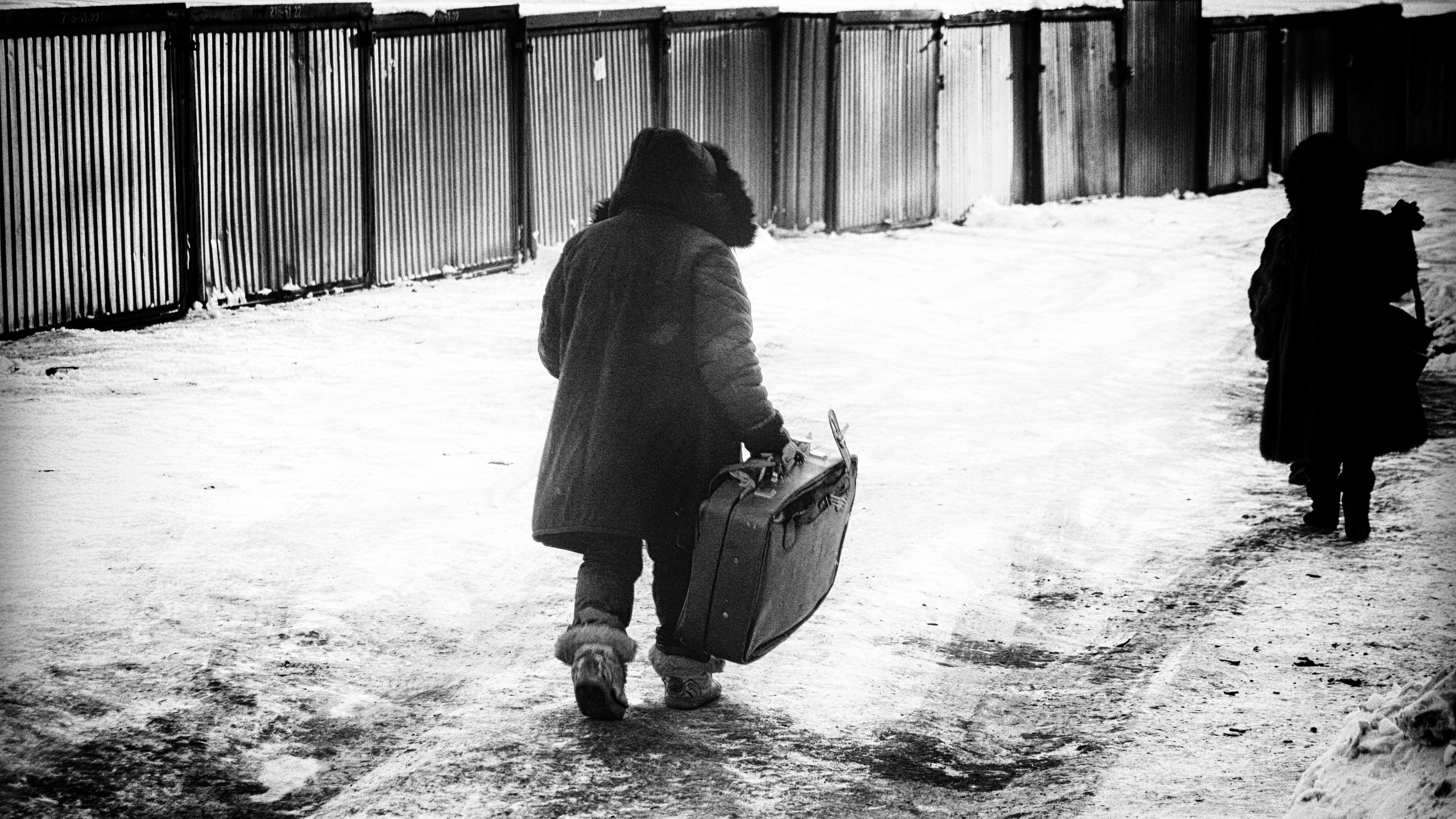 Человек с чемоданом идет по заснеженной дороге мимо гаражей, зимний городской пейзаж, черно-белая уличная фотография о пути