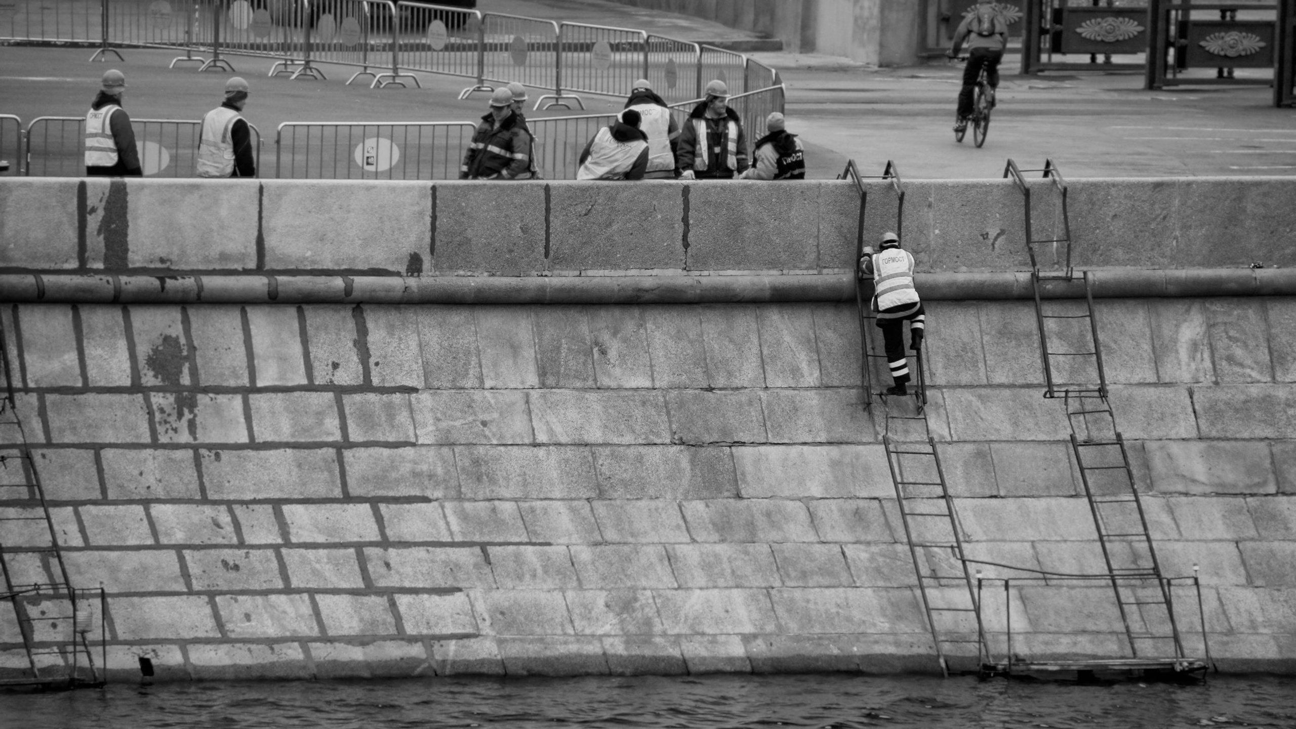 Рабочие в жилетах на каменной набережной с лестницами, велосипедист на фоне, черно-белая уличная фотография