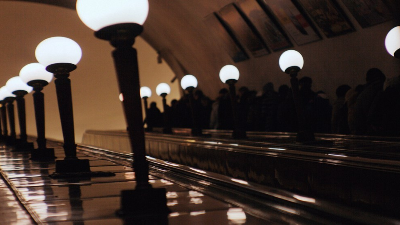 Эскалатор в метро с рядами круглых ламп, силуэты людей наверху, теплый свет, перспектива подземного перехода