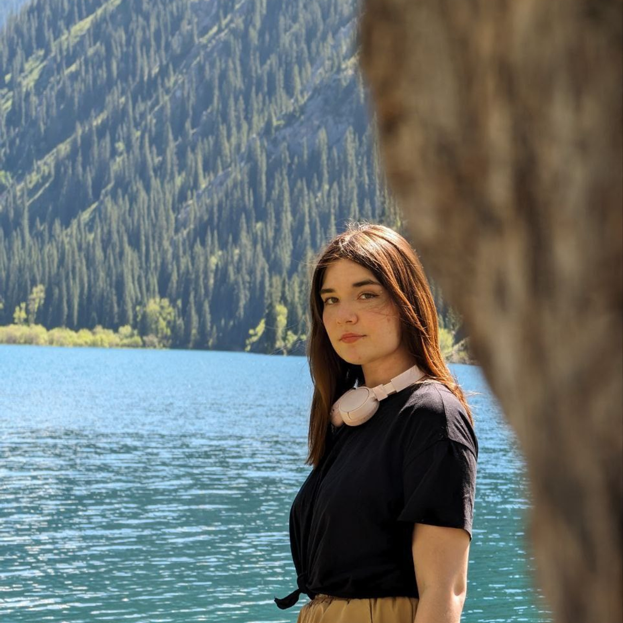 Девушка стоит на фоне живописного горного озера Кольсай с кристально чистой водой, окруженного густым лесом. На ней черная футболка, светло-бежевые брюки и белые наушники на шее. Часть дерева на переднем плане добавляет глубину композиции. Атмосфера спокойная и природная.