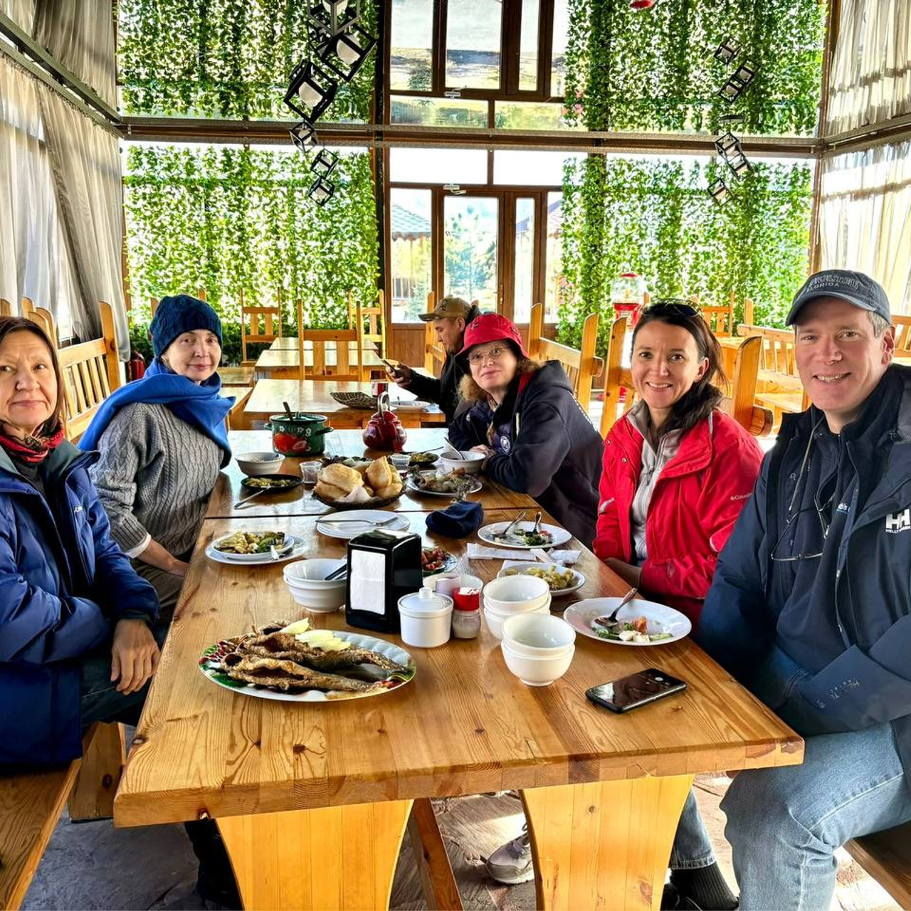 Группа людей сидит за деревянным столом в уютном кафе с панорамными окнами, через которые видны зелёные растения. На столе расположены тарелки с едой, включая жареную рыбу, хлеб и различные блюда. Все участники тепло одеты, некоторые в куртках и шапках, что может свидетельствовать о прохладной погоде. Атмосфера дружеская и непринуждённая.