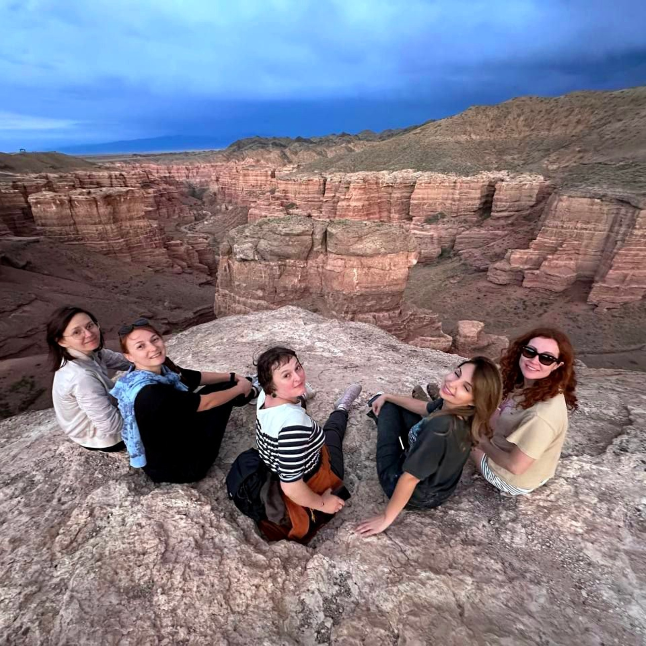 Группа из пяти человек сидит на краю скалы с видом на большой Чарынский каньон с красноватыми стенами. На заднем плане виден драматичный пейзаж с холмами под синим и серым вечерним небом. Люди улыбаются, наслаждаясь видом.