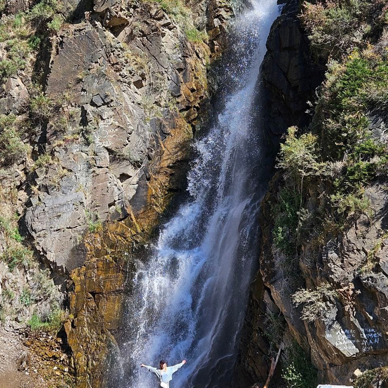 Водопад Медвежий на фоне скал и растительности. Человек стоит у подножия водопада, раскинув руки, создавая ощущение масштаба и величия природы. Вода стекает с высоты, создавая белоснежную пену и поток. На камнях видны яркие природные текстуры, а зелень местами украшает скалы.