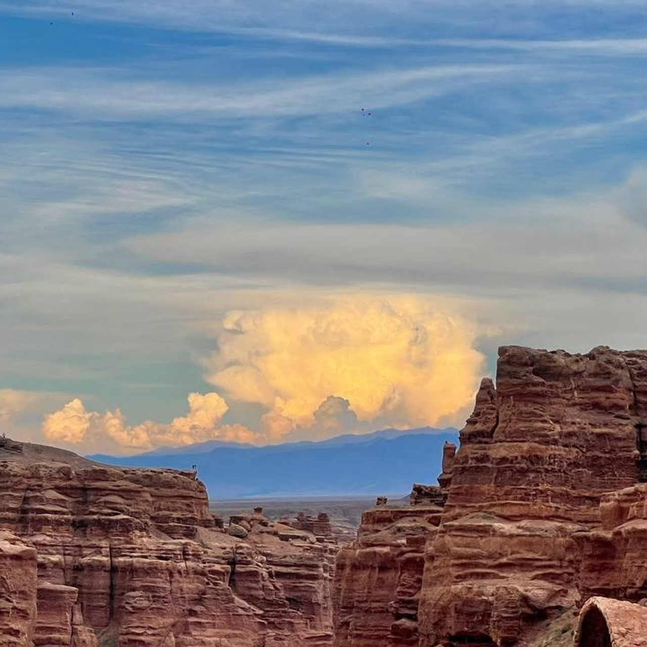Вид на Чарынский каньон с красными скалами под голубым небом. На заднем плане видны ярко-освещённые облака с жёлтым оттенком, за которыми просматриваются далёкие горы. Сочетание цветов каменных стен, облаков и неба создаёт контрастный и живописный пейзаж Долины Замков.