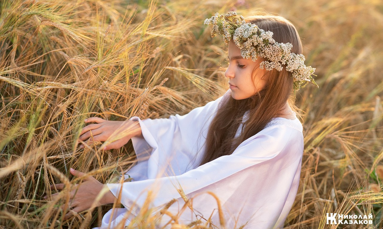 Девушка подросток  с венком на голове и в белом платье трогает колосья пшеницы в лучах закатного солнца.