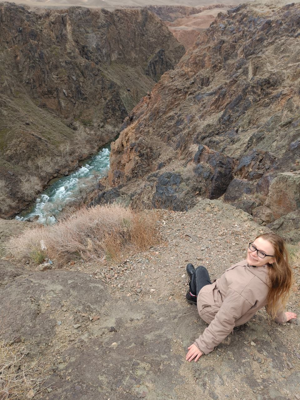 Женщина, путешествующая с компанией Melon Travel, в светло-коричневой куртке сидит на краю утеса с видом на глубокий Черный каньон. Внизу течет извилистая река с бурлящей водой, окруженная крутыми скалистыми стенами. Она улыбается, повернувшись к камере, наслаждаясь моментом и великолепным видом на природу. Ландшафт вокруг сухой, с редкой растительностью.