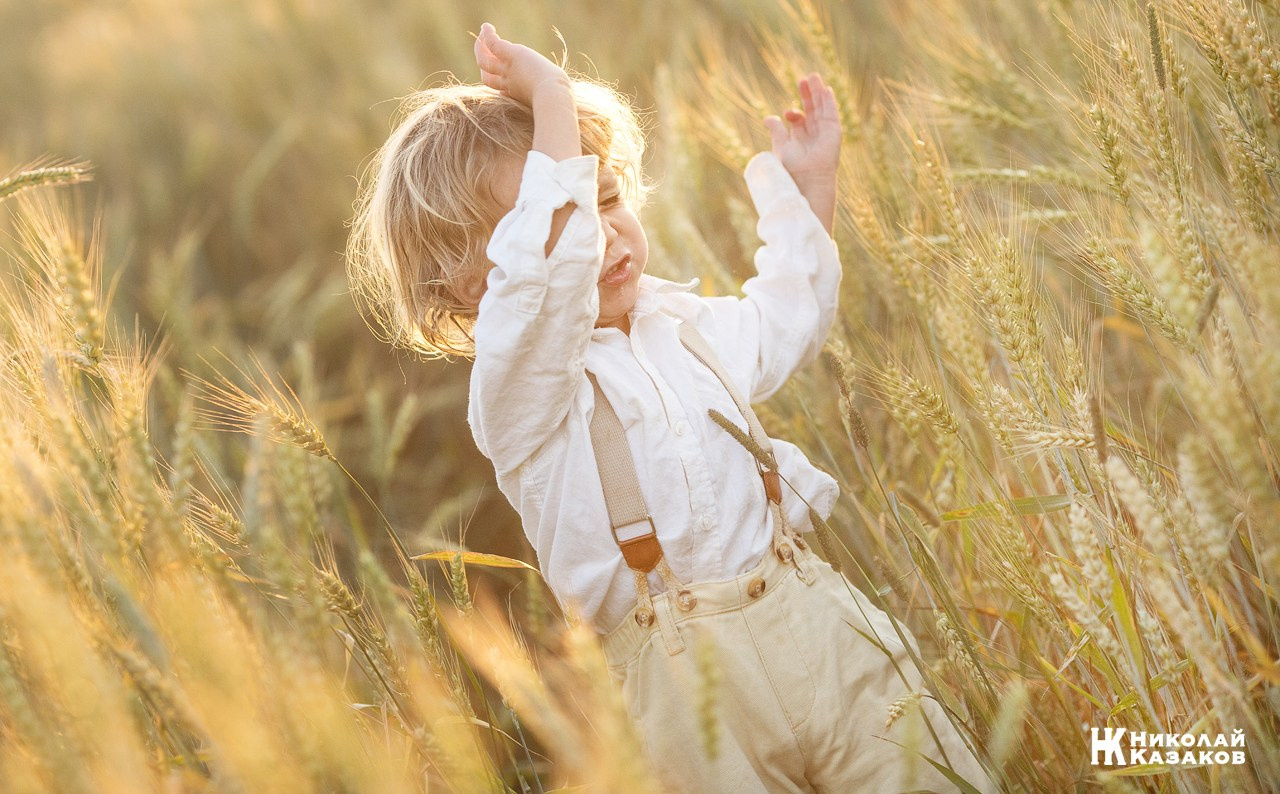 Позитивный портрет маленького мальчика с светлыми волосами в лучах заката, играющий в пшеничном поле во время фотосессии.