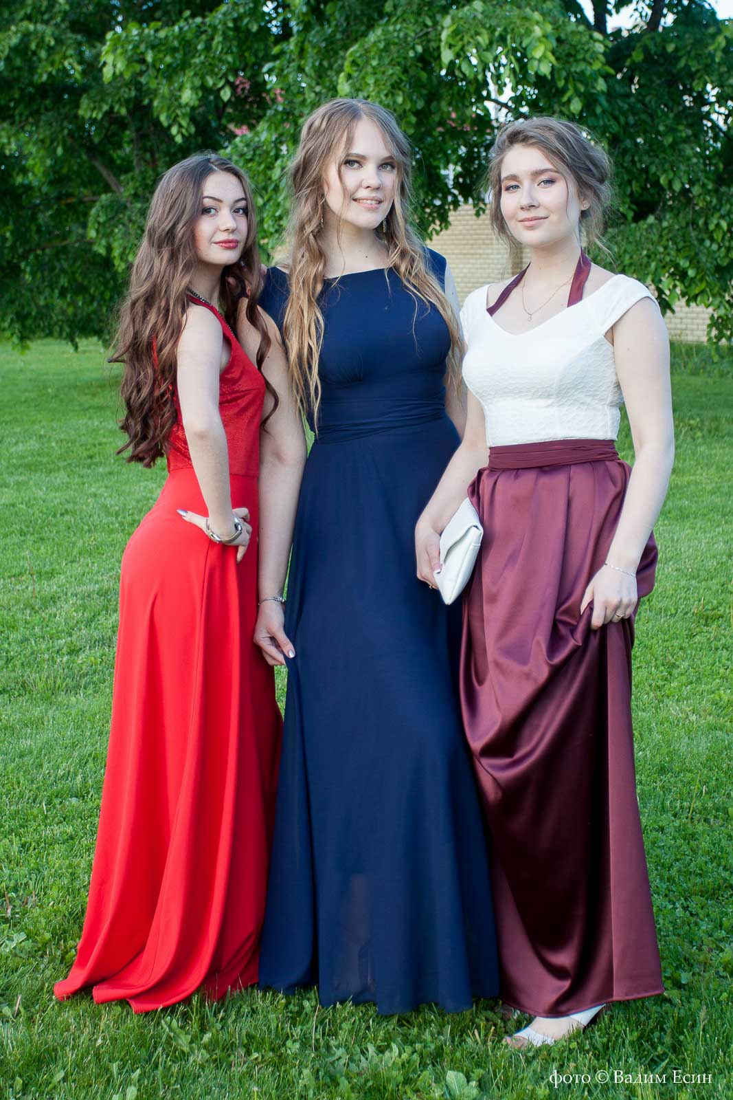 Три девушки с длинными волосами в полный рост стоят на траве.