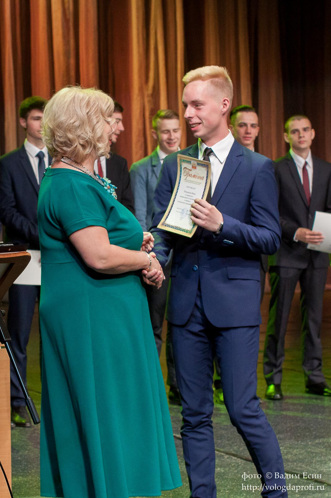 Педагог в зеленом платье торжественно вручает аттестат выпускнику 11 класса в темном синем костюме.