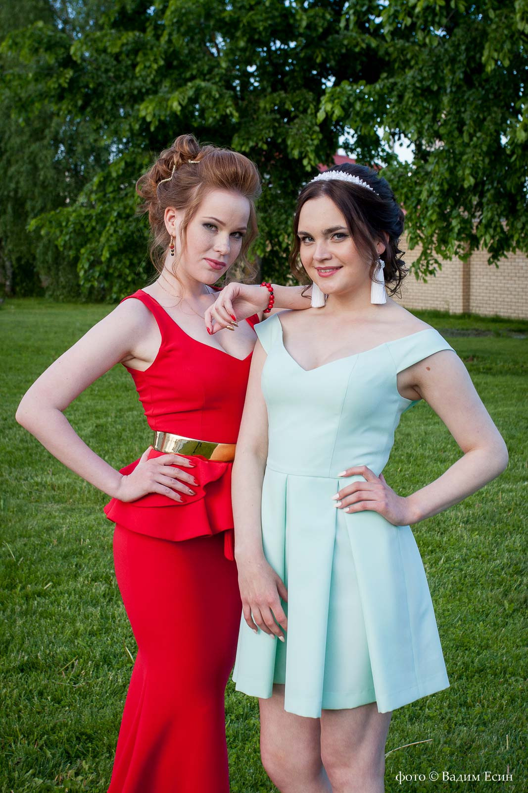Две выпускницы 11 класса в голубом и красном платьях с вечерними прическами стоят рядом и позирую фотографу.