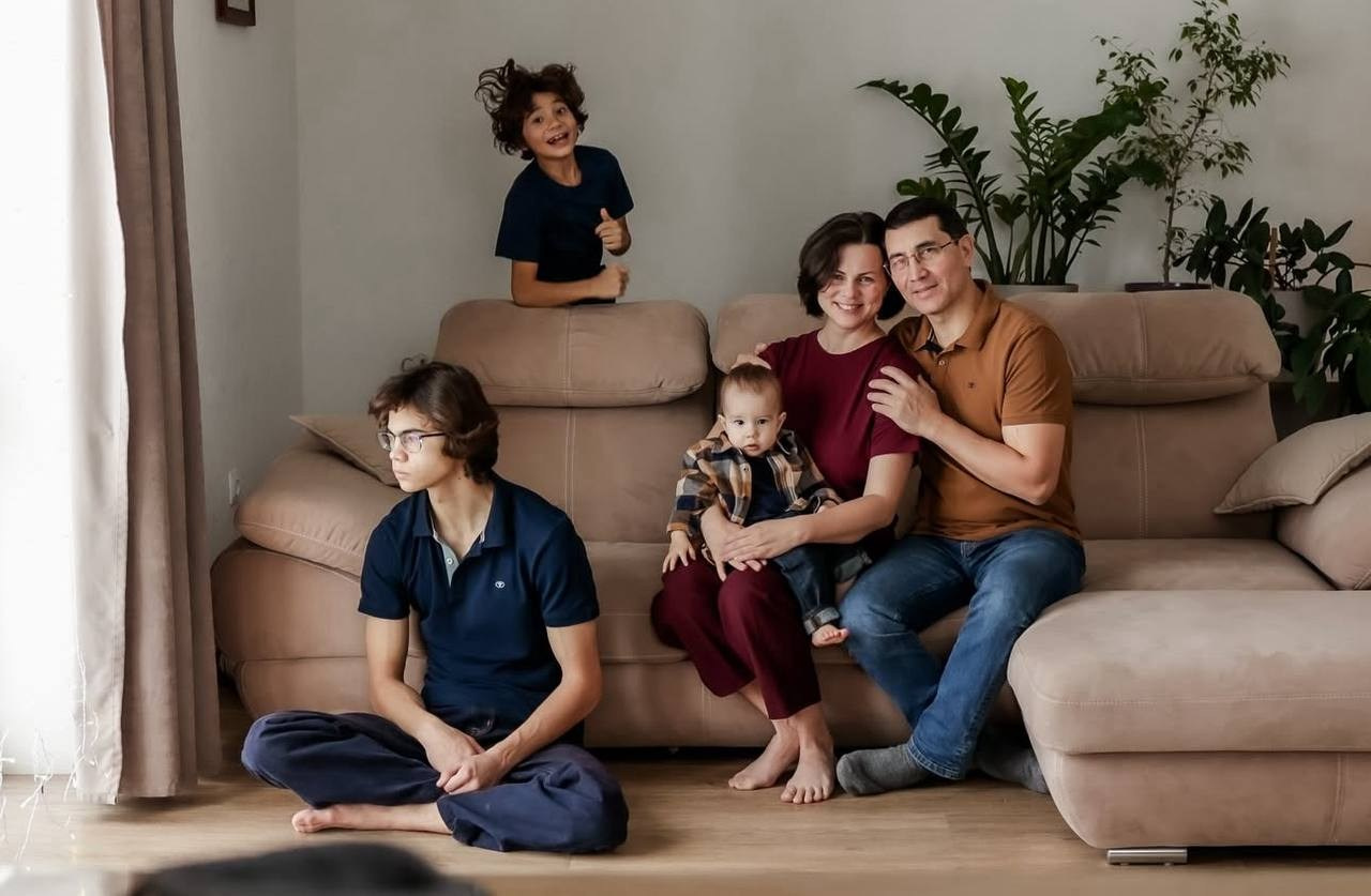 Семья из четырёх человек в светлой гостиной: двое взрослых на диване с малышом, старший ребёнок сидит на полу, ещё один — на спинке дивана.