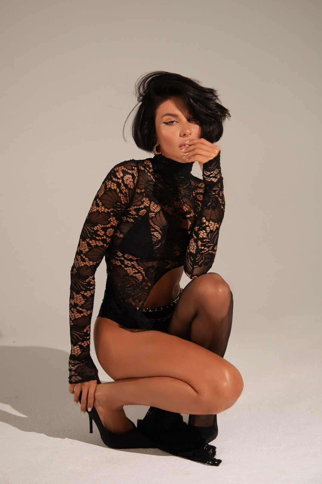 Стильная женщина с короткими темными волосами в черном кружевном боди, колготках и туфлях на каблуке позирует на коленях в студии – модная фотосессия женского белья.