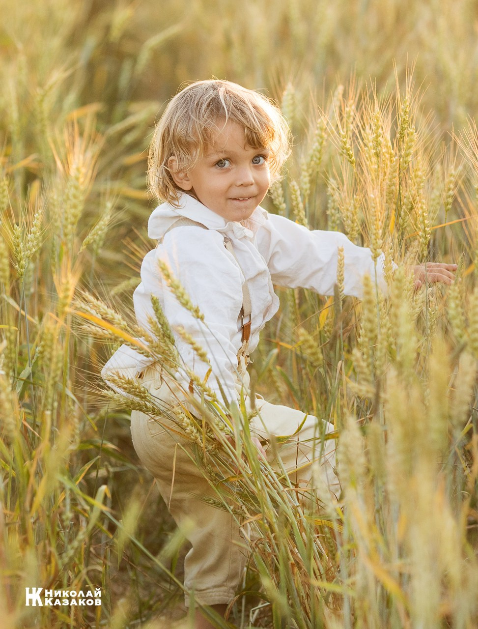 Позитивный портрет маленького мальчика с светлыми волосами в лучах заката, играющий в пшеничном поле во время фотосессии.