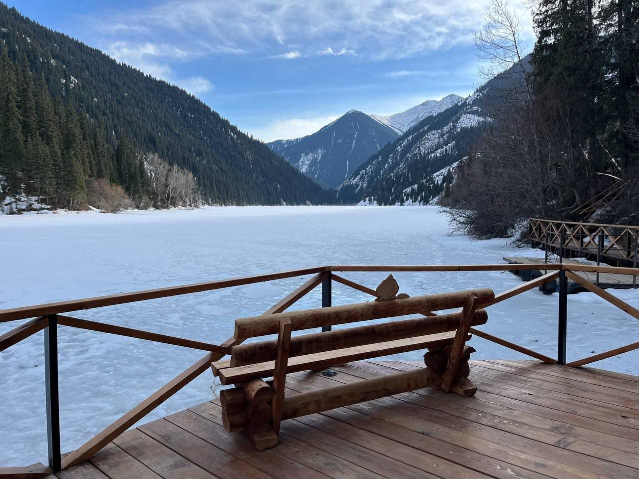 Деревянная скамья на террасе с видом на замёрзшее горное озеро Кольсай рядом с Алматы, окружённое лесистыми склонами и заснеженными горами. Скамья выполнена из резного дерева и расположена в центре деревянной площадки с ограждением. Поверхность озера покрыта снегом и льдом. На заднем плане видны высокие горы, их вершины освещены солнечным светом. Небо ясное, с легкими облаками, создавая атмосферу зимнего уюта и спокойствия.