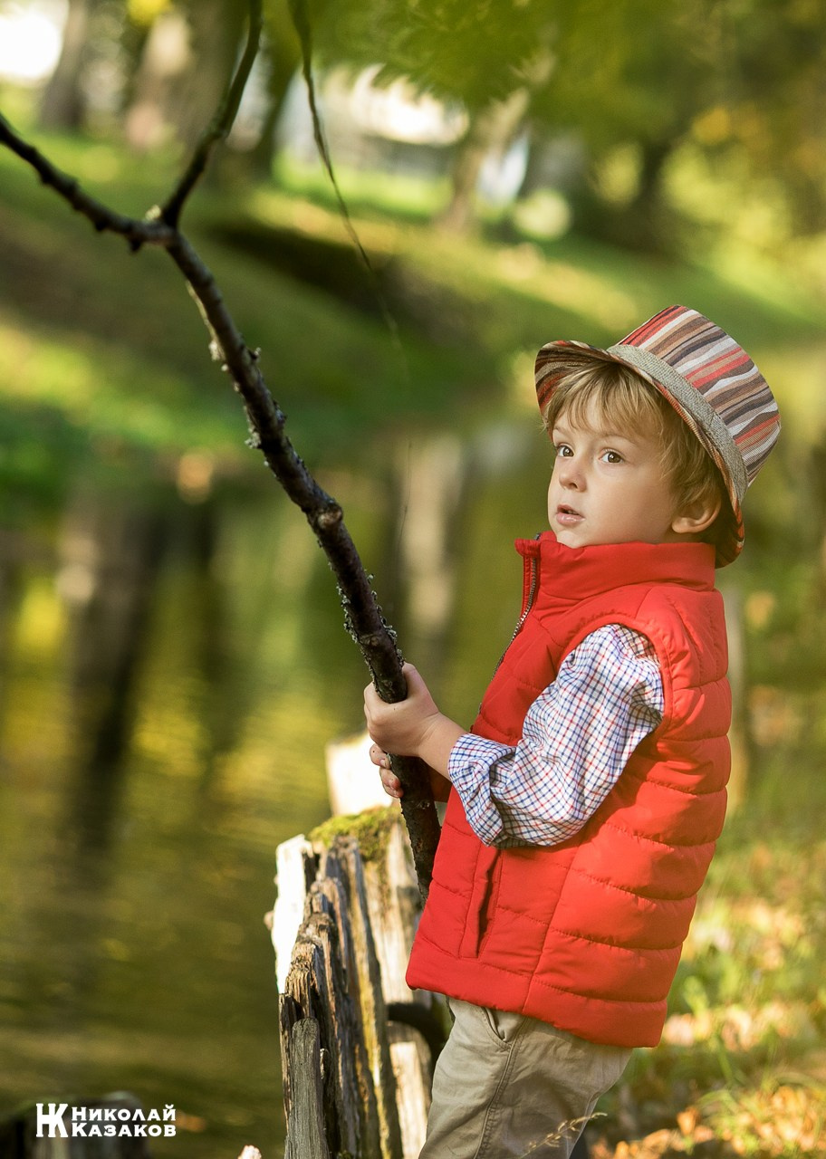 Портрет увлеченного маленького мальчика, ловящего палкой водоросли в речке в лучах утреннего солнца