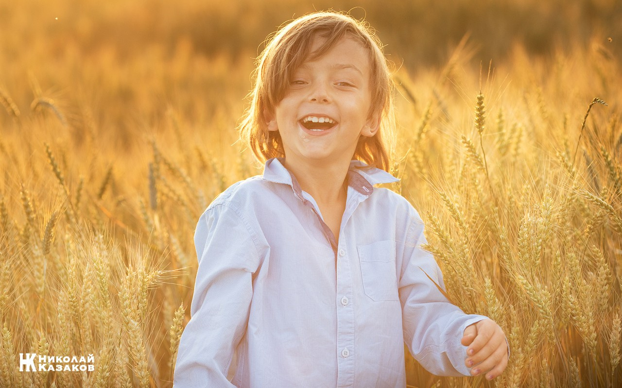 Эмоциональный портрет смеющегося мальчика в лучах закатного солнца, во время фотопрогулки в пшеничном поле на закате.