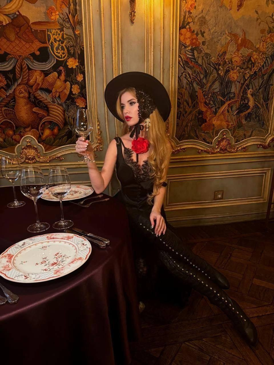 Женщина в черной шляпе с красным цветком на шее сидит за накрытым столом в ресторане