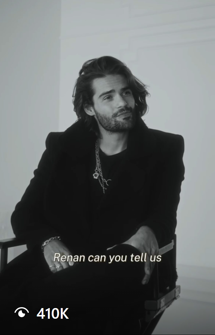 Renan Pacheco giving an interview in black and white, wearing a stylish black coat and layered necklaces, showcasing a modern fashion production house setup.
