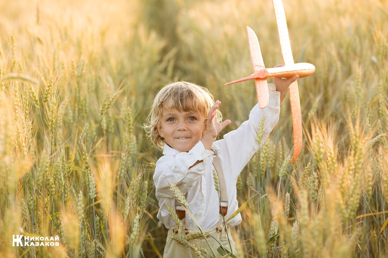 Счастливый маленький мальчик с светлыми волосами идет по пшеничному полу в лучах закатного солнца , держа в руках игрушечный самолет.