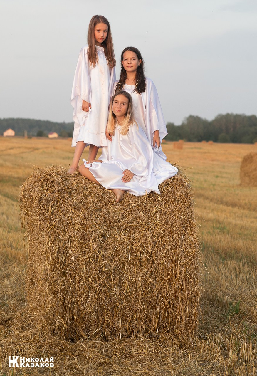 Портрет девочек подростков в белых платьях, стоящих на большом стоге сена во время фотосессии в поле в лучах заката.