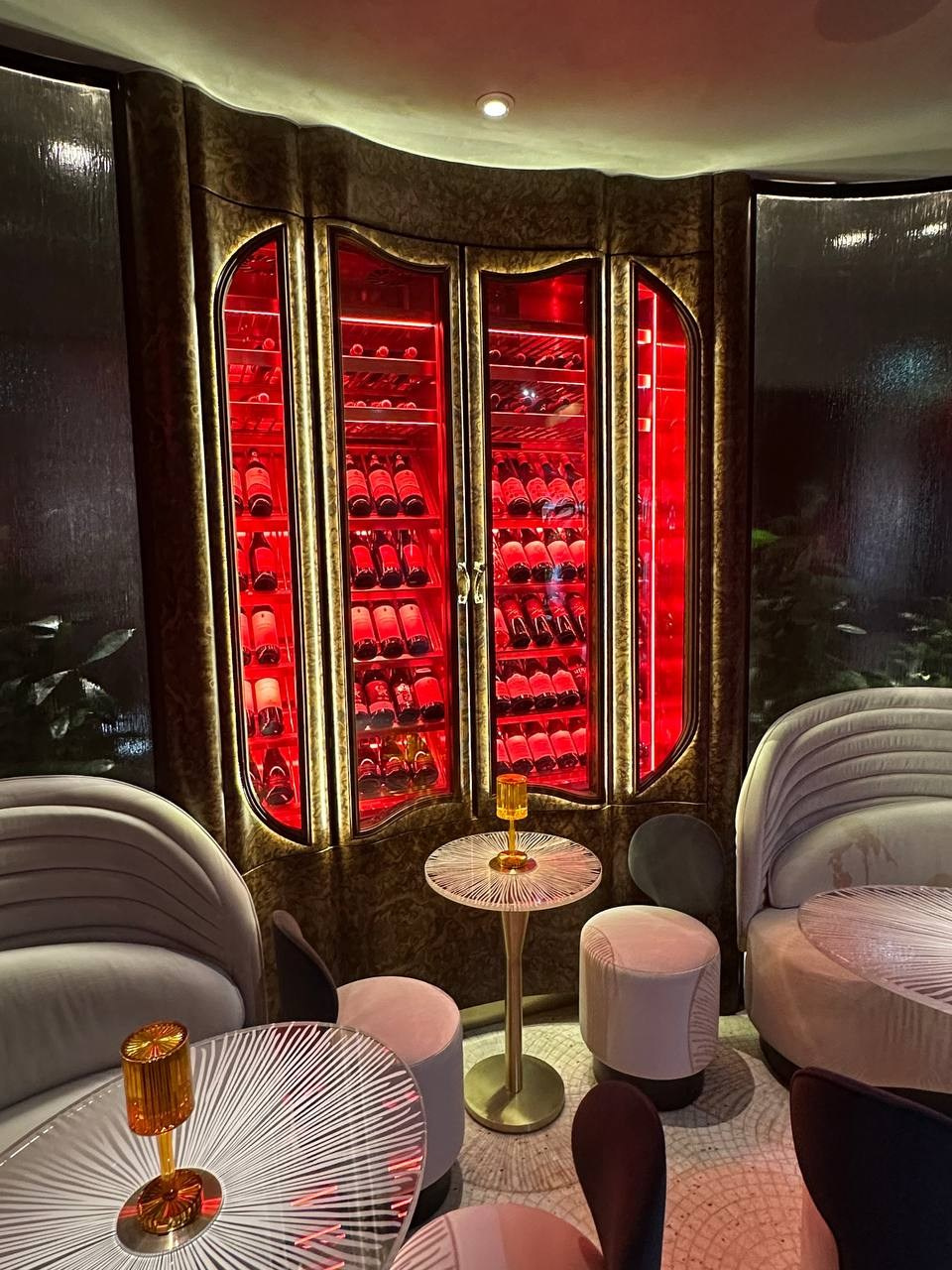 Этот проект родился из сотрудничества с нашими друзьями из компании Winekeys — признанными экспертами в производстве винных шкафов. Они пришли с амбициозной идеей: создать винный шкаф, который станет не просто функциональным элементом ресторана, но произведением искусства. Главный вызов: Облицевать сложные радиусные поверхности шкафа шпоном корня американского ореха — одним из самых капризных и непредсказуемых материалов в столярном деле. Корень ореха славится своей уникальной, почти хаотичной текстурой, где каждый лист шпона неповторим. Процесс создания: Работа началась задолго до первого реза. Десятки встреч с дизайнерами, бесконечные раскладки шпона в поисках идеального сочетания рисунка, множество образцов выкрасов для подбора финишного покрытия. Каждый элемент требовал индивидуального подхода. Техника раскладки: Мы использовали принцип «книжки» — классический прием, когда листы шпона раскрываются как страницы книги, создавая зеркально-симметричный рисунок. Это позволило проявить знаменитые «морды» — характерные завихрения текстуры корня американского ореха, которые образуют почти портретные, живые узоры. Сложность радиусных форм требовала не только технического мастерства, но и художественного видения — каждая деталь должна была встать на свое место так, чтобы общая композиция читалась как единое целое, несмотря на непредсказуемость природного материала. Результат — монументальный винный шкаф, где благородство корня ореха подчеркивает ценность каждой бутылки внутри.