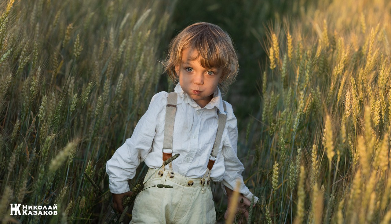 Позитивный портрет маленького мальчика с светлыми волосами в лучах заката, играющий в пшеничном поле во время фотосессии.
