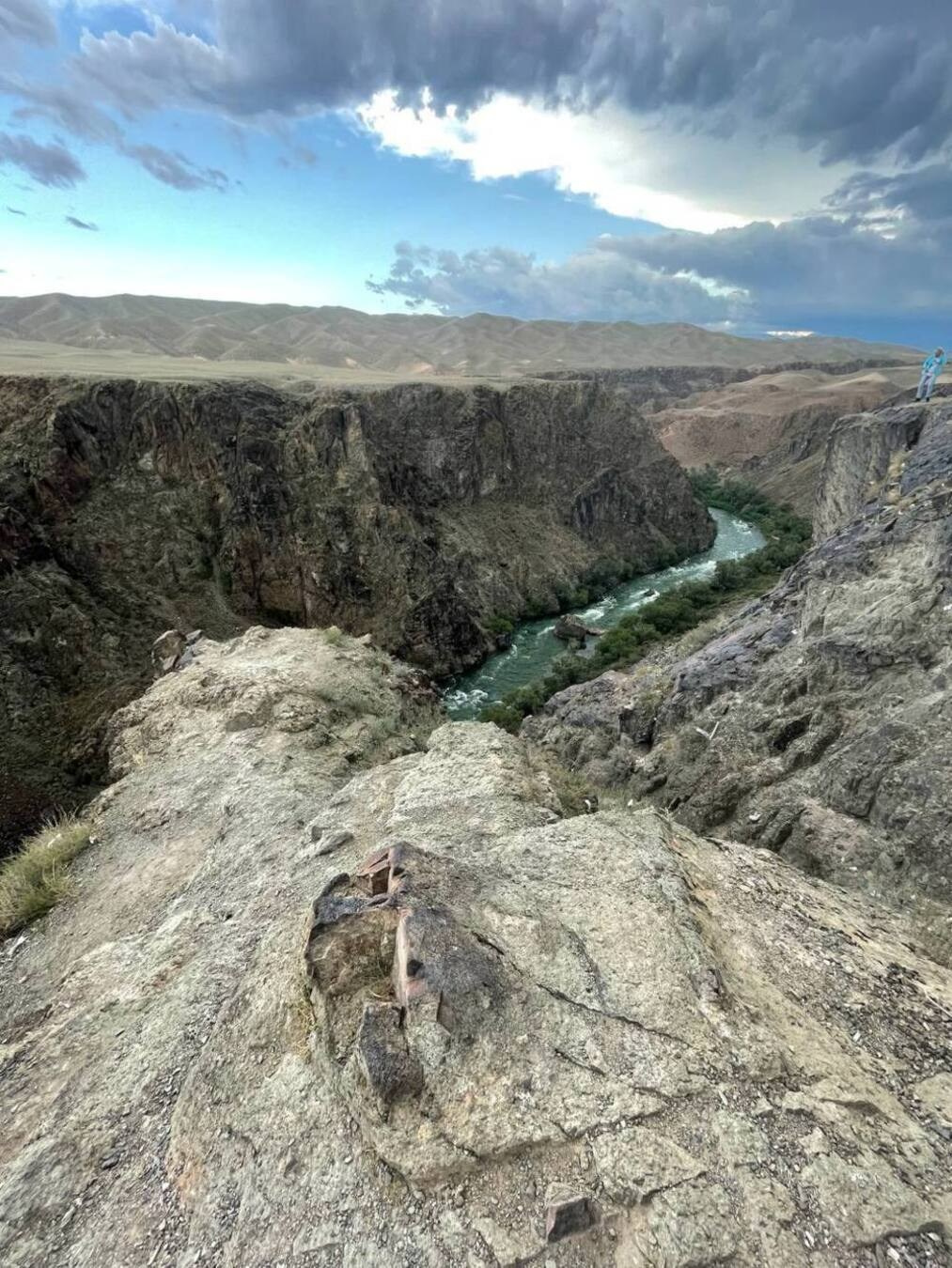 Живописный вид на глубокий Черный каньон с извилистой рекой внизу. Берега каньона покрыты зеленью, а окружающий ландшафт представляет собой сухие холмы и скалы. Небо затянуто облаками, создавая драматичную атмосферу, но на горизонте виднеются просветы голубого неба. На переднем плане крупный выступ скалы с грубой текстурой, а справа вдалеке человек стоит на краю обрыва, подчеркивая величие природы.