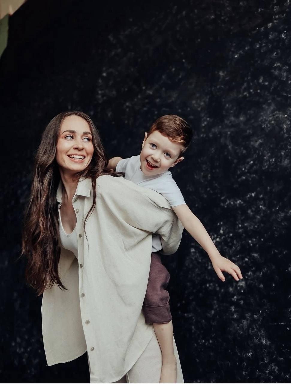 Мама с улыбкой несёт на спине смеющегося сына. Тёмный фон, атмосфера весёлая, тёплая, игривая.