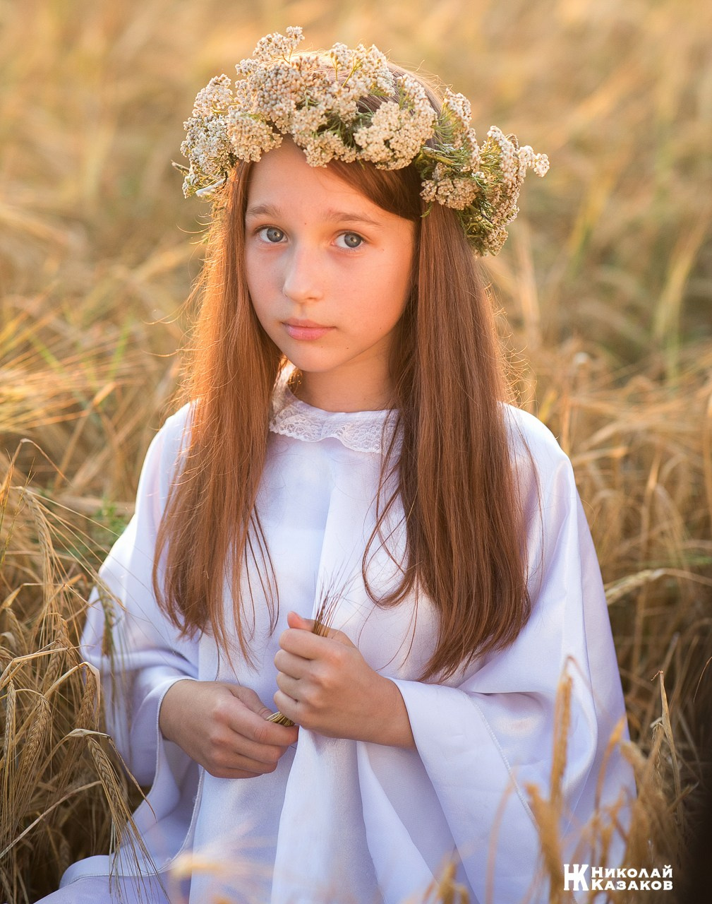 Эмоциональный портрет нежной девочки с венком на голове в лучах закатного солнца , сидящая в колосьях пшеницы во время фотосессии.