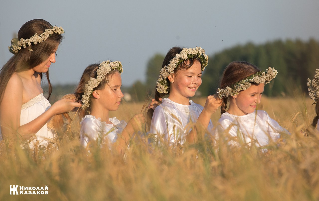 Танцевальная группа девочек подростков  с венками на головах плетут косы сидя в пшенчном поле на закате во время фотосессии