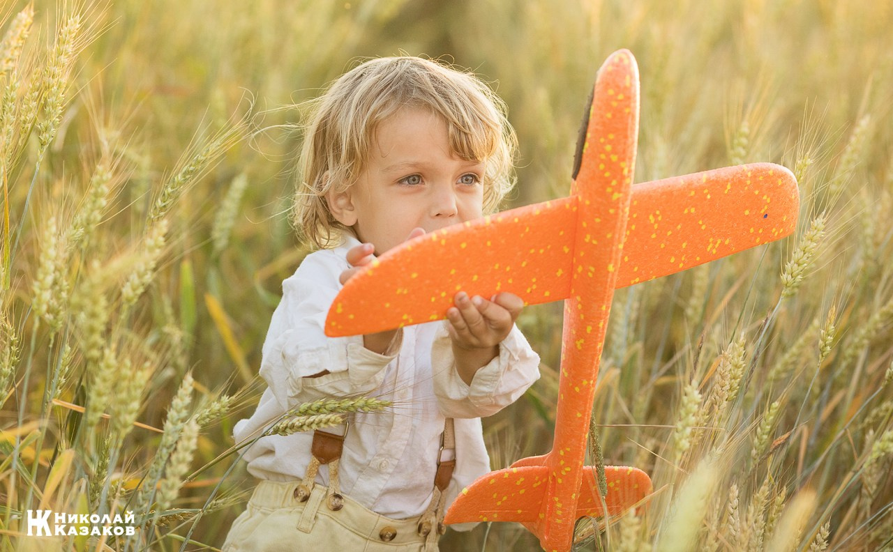Счастливый маленький мальчик с светлыми волосами резвится в пшеничном поле в лучах закатного солнца , играя с игрушечным самолетом.