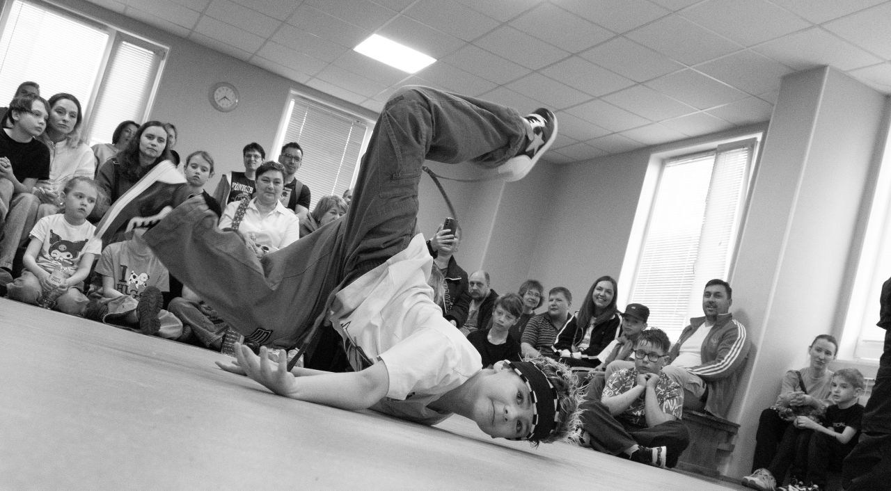 Чёрно-белое фото: молодой брейк-дансер выполняет фриз (прогиб на груди) на полу в помещении, вокруг сидят зрители разных возрастов; документальная съёмка танцевального выступления или баттла