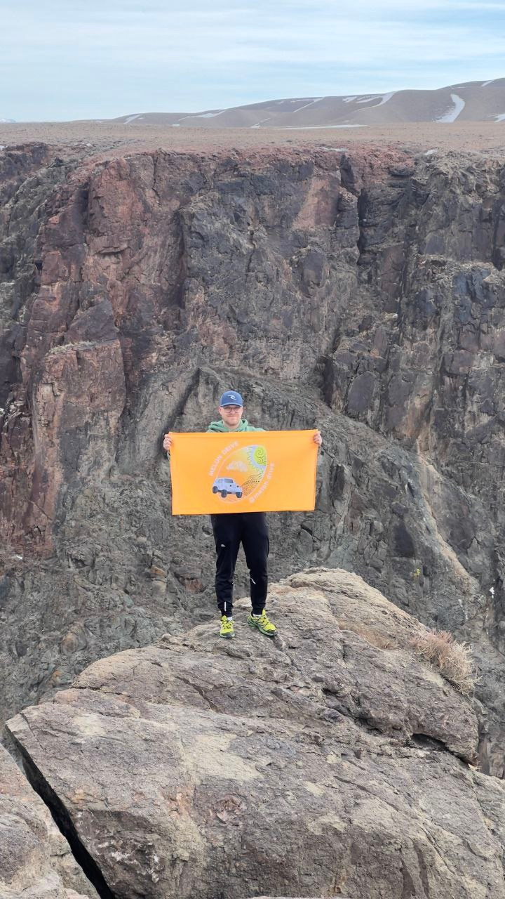 Мужчина стоит на фоне Черного каньона и держит в руках оранжевый флаг с логотипом туристического агентства Melon Drive и рисунками, на фоне которого изображены автомобиль и природный пейзаж. Он одет в спортивную одежду: черные брюки, зелёную кофту и ярко-жёлтые кроссовки. На голове кепка. На заднем плане видно высокие отвесные скалы и холмы, переходящие в равнину под облачным небом.