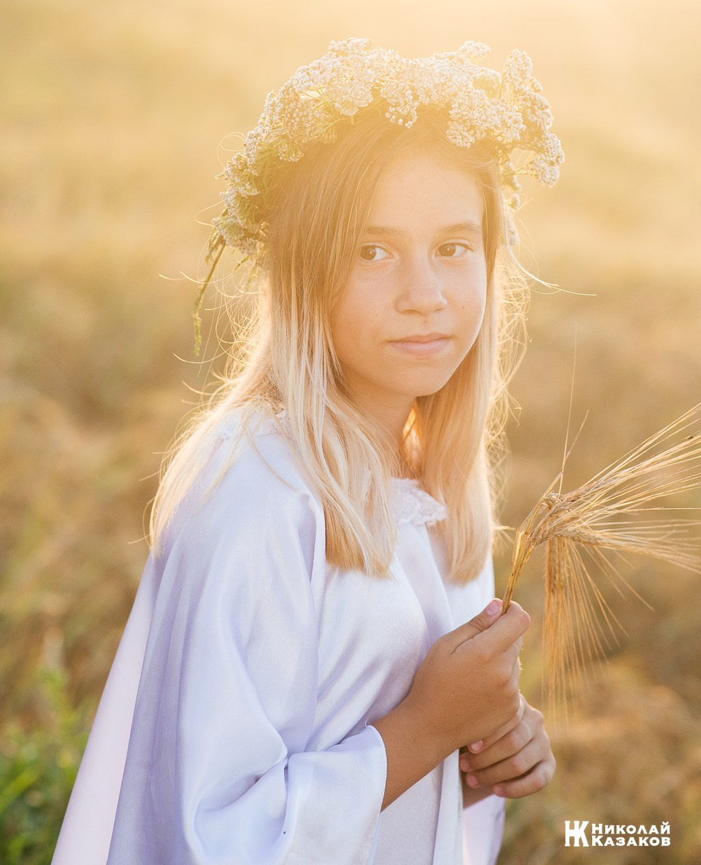 Портрет счастливой девочки с венком на голове в лучах закатного солнца во время фотосессии в пшеничном поле.