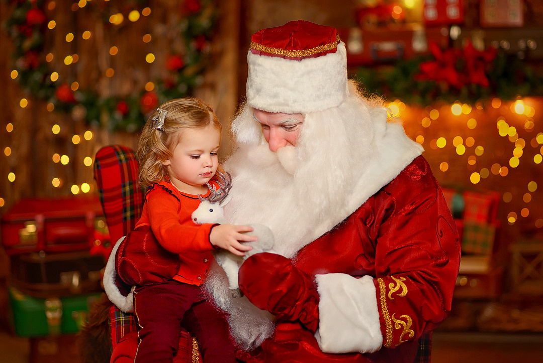 Дед Мороз в Новогодней студии дарит маленькой девочке в подарок игрушечного оленёнка.