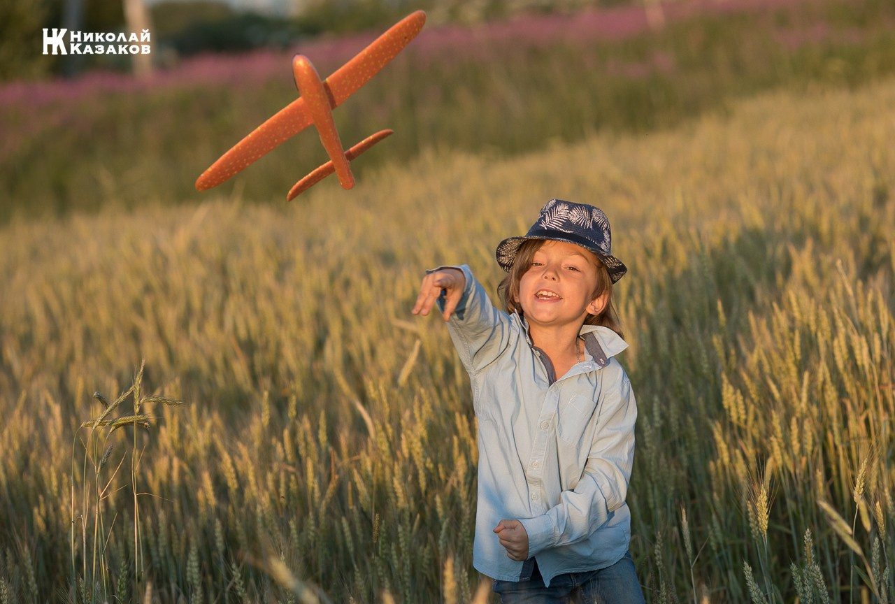 Счастливый маленький мальчик с светлыми волосами играет в пшеничном поле в лучах закатного солнца , запуская игрушечный самолет.